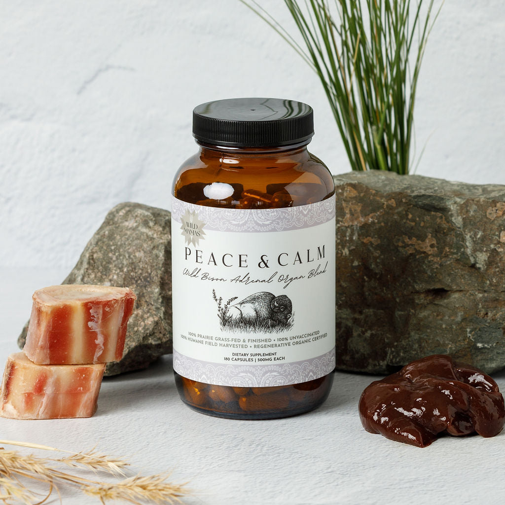 A brown glass jar labeled "Peace and Calm- Wild Bison Adrenal Organ Blend" by Wild Mamas sits on a white surface with rocks, raw meat, and grass. The label displays a sleeping animal and calming, natural colors.