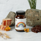 A bottle of Wild Mamas Wild Child - Complete Multivitamin For Kids sits on a surface with stacked bones, yellow flowers, grass, brown paste, and rocks against a light grey background—featuring a bison organ blend as an early food for babies.