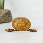 The Wild Mamas Shilajit + Decaffeinated Coffee Bean Deeply Exfoliating Bison Tallow Soap Scrub stands upright on a white surface with scattered coffee beans, set against a blurred rock and green plant to emphasize its organic, textured ingredients.