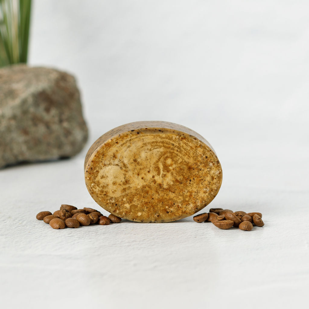 The Wild Mamas Shilajit + Decaffeinated Coffee Bean Deeply Exfoliating Bison Tallow Soap Scrub stands upright on a white surface with scattered coffee beans, set against a blurred rock and green plant to emphasize its organic, textured ingredients.