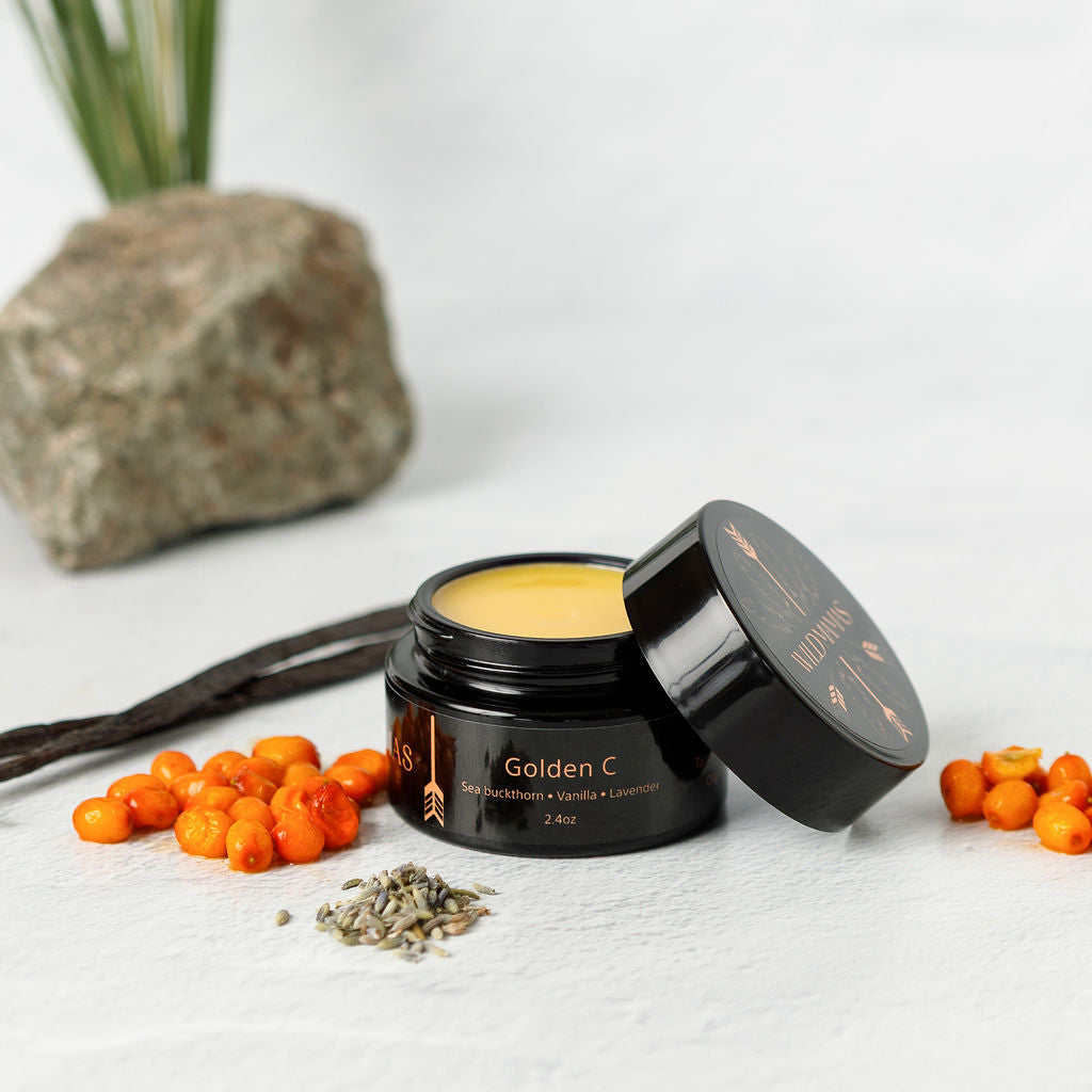 A sleek black jar with gold text of Wild Mamas Golden C – Wild Bison Tallow Balm sits open on a white surface, glowing yellow and surrounded by sea buckthorn berries, lavender buds, and vanilla beans for a brightening vitamin C boost.