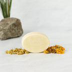 A pale yellow oval bar of Wild Mamas Chamomile + Calendula (Baby)- Wild Bison Tallow Soap rests on a white surface, surrounded by dried chamomile and calendula flowers, with a rock and green leaves in the background for a soothing effect.