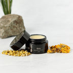 A black jar of Wild Mamas Baby- Wild Bison Tallow Balm with its lid off sits on a white surface, surrounded by dried chamomile and calendula flowers, a rock, and a plant in the background.