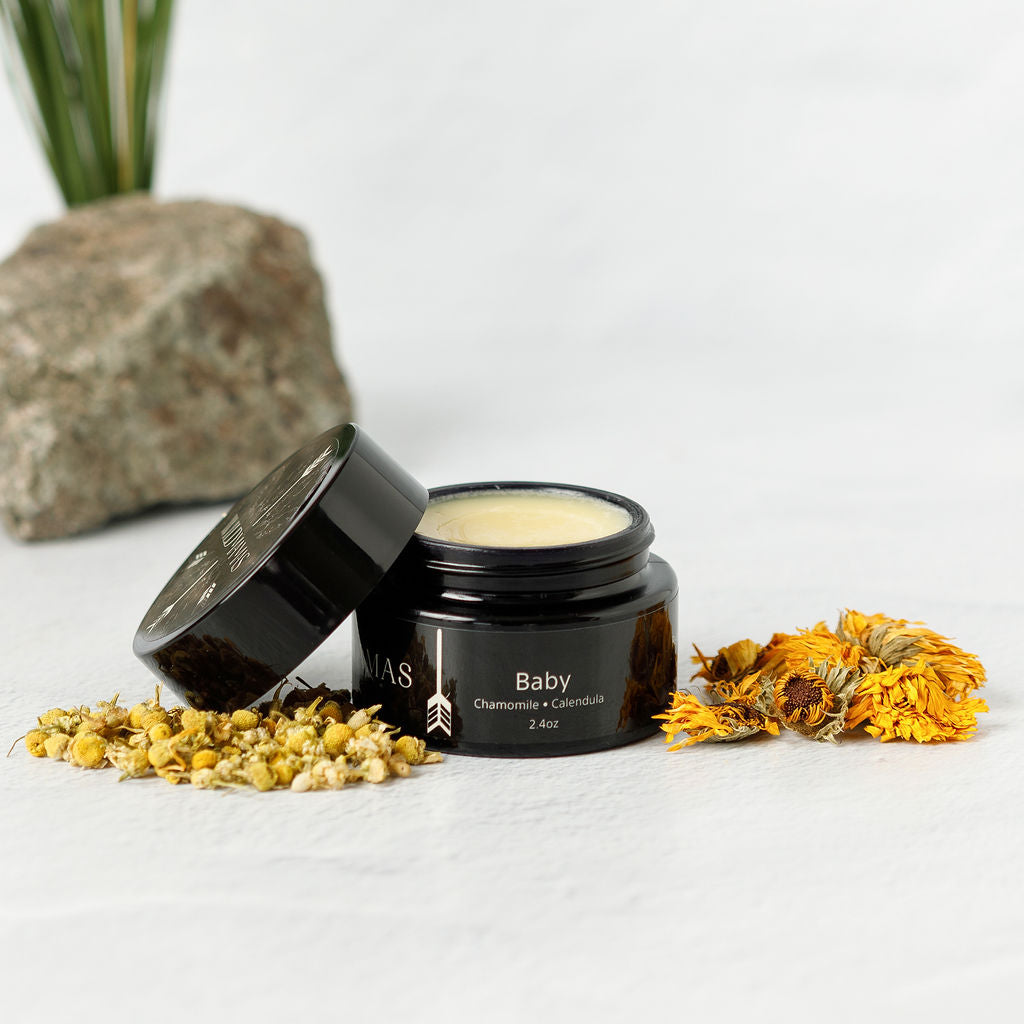 A black jar of Wild Mamas Baby- Wild Bison Tallow Balm with its lid off sits on a white surface, surrounded by dried chamomile and calendula flowers, a rock, and a plant in the background.