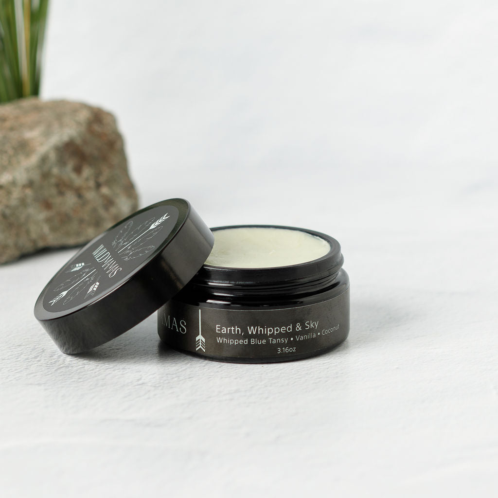 A black jar of Wild Mamas Earth, Whipped & Sky 3.16oz Wild Bison Tallow Balm (Whipped) sits on a white surface with the lid partly off, showing creamy tallow inside; a rock and green plant are in the background.