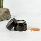 A black jar of Wild Mamas Ancestral Blue 2.4oz Wild Bison Tallow Balm sits open, revealing cream inside. Next to it lie yellow powder and glossy orange liquid on a light surface, with a rock and green plant in the background.