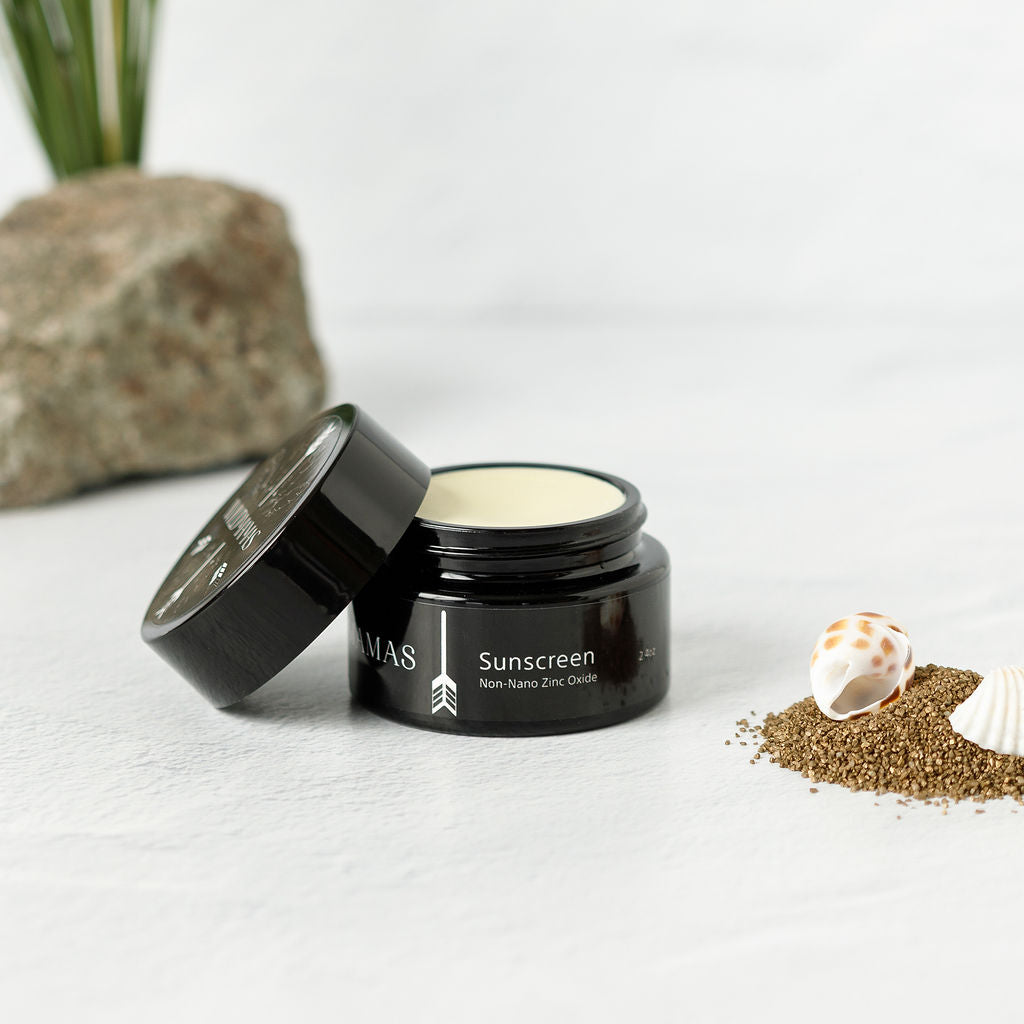 A black jar of Wild Mamas Sunscreen- Wild Bison Tallow Balm with the lid off sits on a white surface, beside sand, two seashells, and a rock, with greenery in the background.