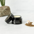 A black jar of Wild Mamas Sunscreen- Wild Bison Tallow Balm with the lid off sits on a white surface, beside sand, two seashells, and a rock, with greenery in the background.