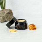 An open black jar of Wild Mamas' Honey Bee (Raw Honey) - Wild Bison Tallow Balm sits next to beeswax pellets and honeycomb, with a rock and green plant in the background. The creamy balm features regenerative organic skincare ingredients.