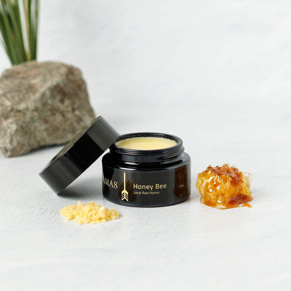 An open black jar of Wild Mamas' Honey Bee (Raw Honey) - Wild Bison Tallow Balm sits next to beeswax pellets and honeycomb, with a rock and green plant in the background. The creamy balm features regenerative organic skincare ingredients.