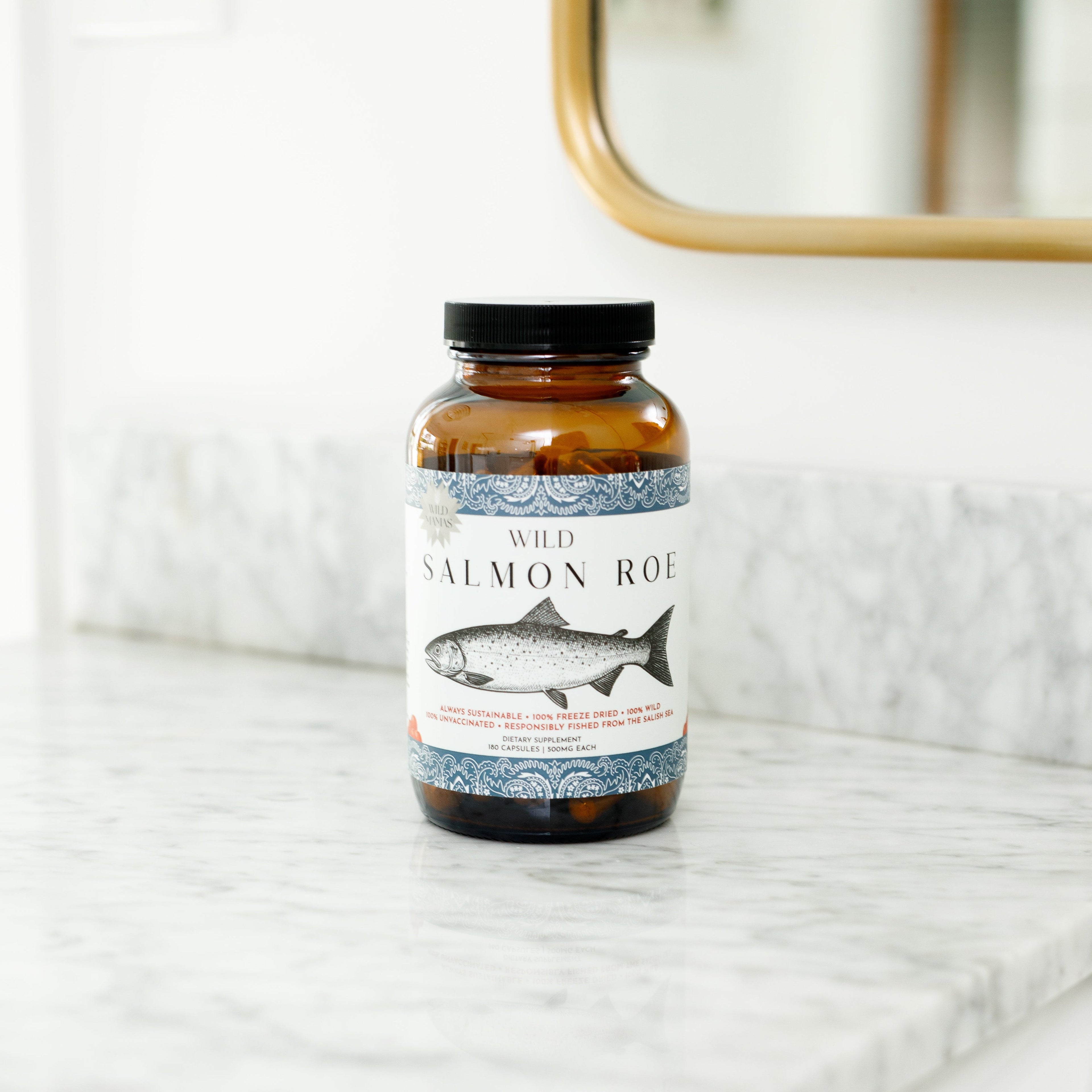A glass jar of Wild Mamas Wild Salmon Roe, featuring a fish illustration, sits on a marble countertop in a bright room with part of a mirror visible in the background, emphasizing whole-food nutrition.