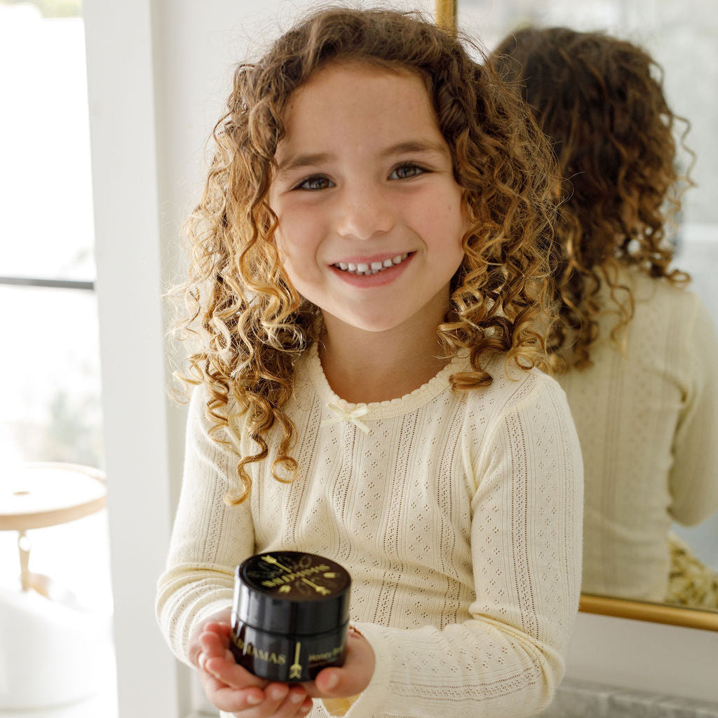 A smiling young child with curly hair holds a jar of Wild Mamas Honey Bee (Raw Honey)-Wild Bison Tallow Balm while wearing a cream long-sleeve top, standing in front of a mirror.