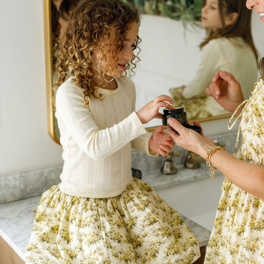 A young girl in a cream and yellow floral dress sits on a bathroom counter, smiling as she dips her finger into Wild Mamas Baby - Wild Bison Tallow Balm held by an adult, with their reflection visible in the mirror behind them.