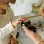 A smiling child dips a finger into a jar of Wild Mamas Baby- Wild Bison Tallow Balm held by an adult near a bathroom sink, both dressed in cream-colored clothes.
