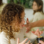 A young girl in a cream top has Wild Mamas Honey Bee (Raw Honey)-Wild Bison Tallow Balm gently dabbed on her face by an adult, with her reflection visible in the background mirror.