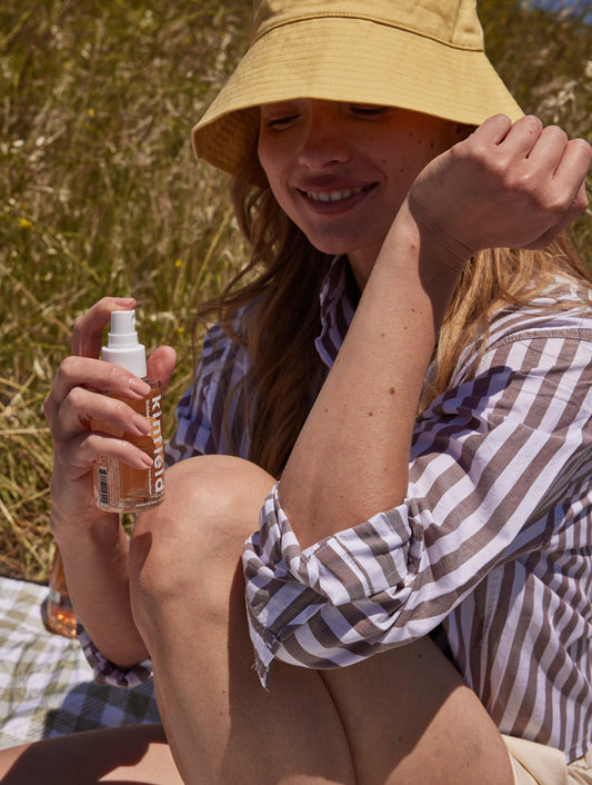 A person wearing a yellow bucket hat and striped shirt sits outdoors on a picnic blanket, smiling while holding a spray bottle labeled “Golden Hour™” from Kinfield. Sunlight filters through the grassy area, highlighting the clean bug spray made with Indonesian citronella.