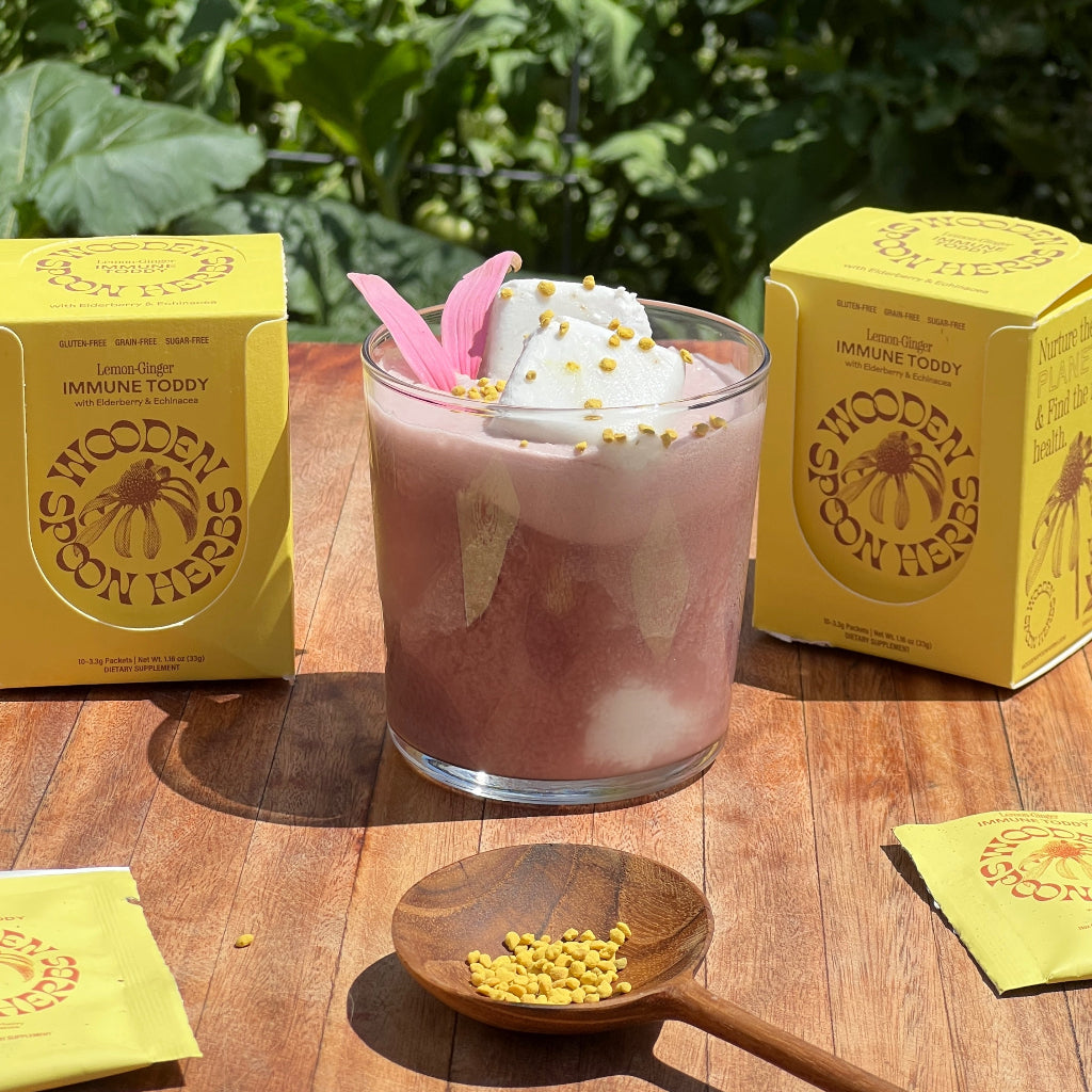 A glass of pink drink with ice, whipped topping, a pink flower, and yellow sprinkles sits on a wooden table next to two WOODEN SPOON HERBS Lemon-Ginger Immune Toddy boxes. Green plants can be seen in the background.