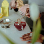 Someone stirs a cup of WOODEN SPOON HERBS Lemon-Ginger Immune Toddy with a wooden spoon, while ginger is grated nearby among tea ingredients and elderberry, using a silver dish for natural immune support.