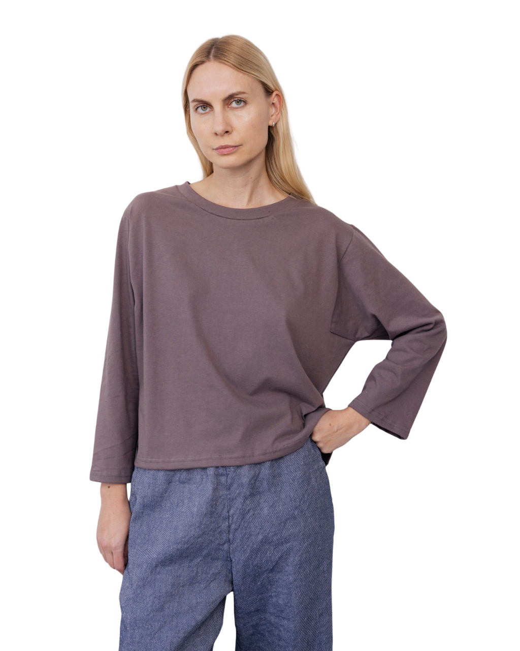 A woman with straight blonde hair wears the Wol Hide Long Sleeve Tee in Mushroom and blue pants, standing against a plain white background with one hand on her hip.