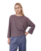 A woman with straight blonde hair wears the Wol Hide Long Sleeve Tee in Mushroom and blue pants, standing against a plain white background with one hand on her hip.