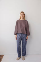 A woman with long blonde hair stands indoors against a white wall, wearing the Wol Hide Long Sleeve Tee in Mushroom, blue-gray pants, and beige shoes. She has a neutral expression and her arms relax at her sides.