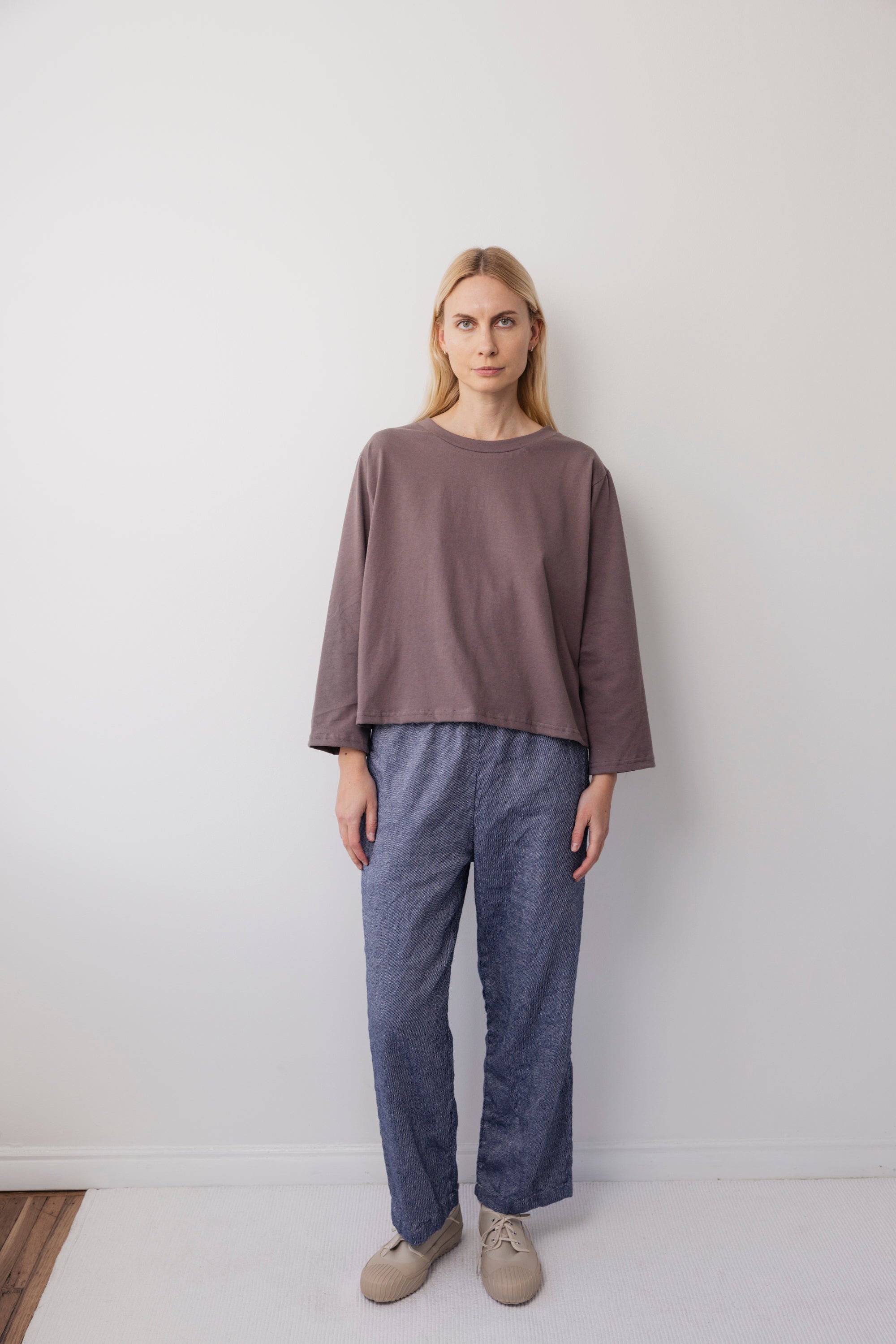 A woman with long blonde hair stands indoors against a white wall, wearing the Wol Hide Long Sleeve Tee in Mushroom, blue-gray pants, and beige shoes. She has a neutral expression and her arms relax at her sides.