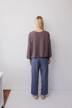 A person with long blond hair faces a white wall, wearing Wol Hide’s Long Sleeve Tee in Mushroom and blue wide-leg pants. Their beige shoes are on a light floor, creating a minimalistic scene.