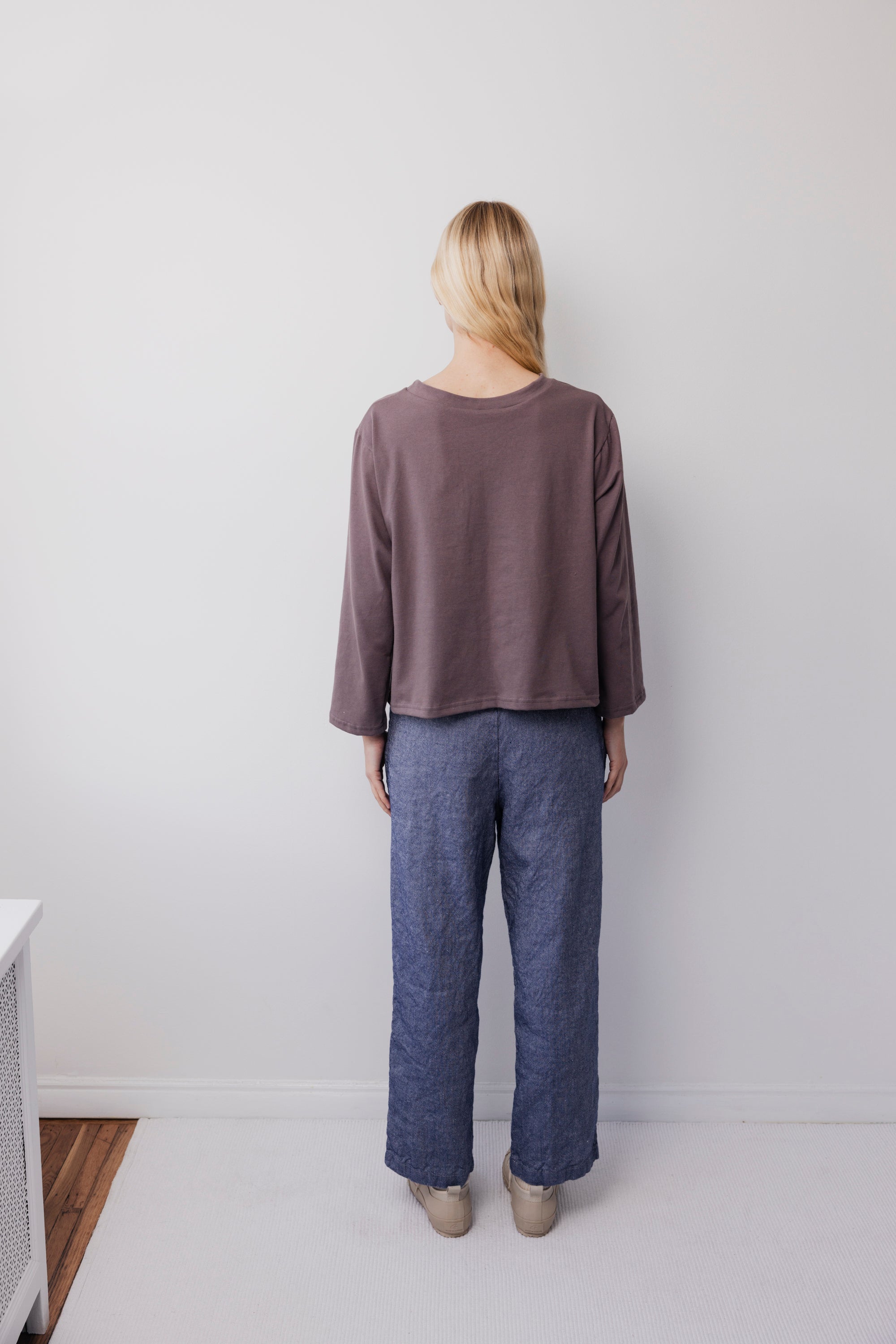 A person with long blond hair faces a white wall, wearing Wol Hide’s Long Sleeve Tee in Mushroom and blue wide-leg pants. Their beige shoes are on a light floor, creating a minimalistic scene.
