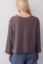 A person with long blonde hair faces away from the camera against a plain light background, wearing Wol Hide’s Long Sleeve Tee in Mushroom and blue pants.