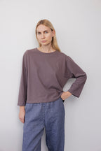 A woman with long blonde hair stands against a plain light wall, wearing the Wol Hide Long Sleeve Tee in Mushroom and blue pants, one hand on her hip with a neutral expression.