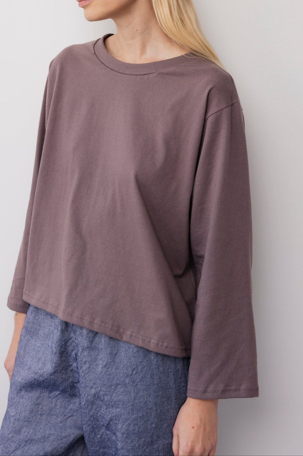 A woman wears the Wol Hide Long Sleeve Tee in Mushroom, a loose-fitting organic cotton top with wide sleeves, paired with high-waisted blue pants. She stands against a light wall, her face partially out of frame.