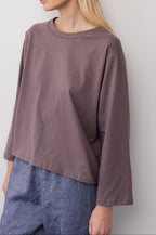A woman wears the Wol Hide Long Sleeve Tee in Mushroom, a loose-fitting organic cotton top with wide sleeves, paired with high-waisted blue pants. She stands against a light wall, her face partially out of frame.