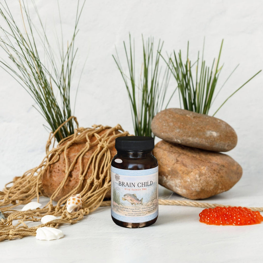 A bottle of Wild Mamas Brain Child rests on a fishing net with seashells, wild salmon roe, rocks, and tall grass against a white background—evoking the natural essence of baby’s first food.