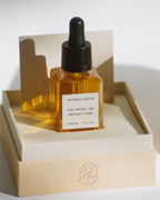 A square glass dropper bottle labeled "THE FACIAL OIL" by THOMAS GROVE SKIN rests in an open cream box with branded lid, on a white surface in soft natural light—ideal for organic skincare and anti-aging routines.