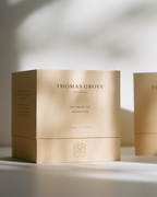 A beige box labeled "THOMAS GROVE SKIN" and "THE FACIAL OIL" stands on a white surface with soft shadows and sunlight, showcasing the elegant, minimalistic packaging of this premium organic skincare product.