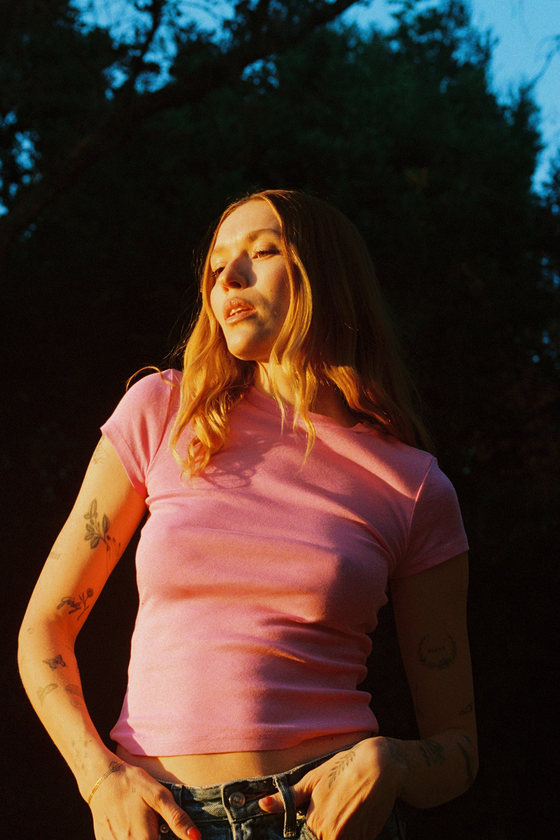 A person with long hair stands outdoors under the setting sun, wearing a pink LA Relaxed Bebe Organic Tee and denim jeans. Their tattooed arms are highlighted by warm lighting, with trees and a clear sky in the background.