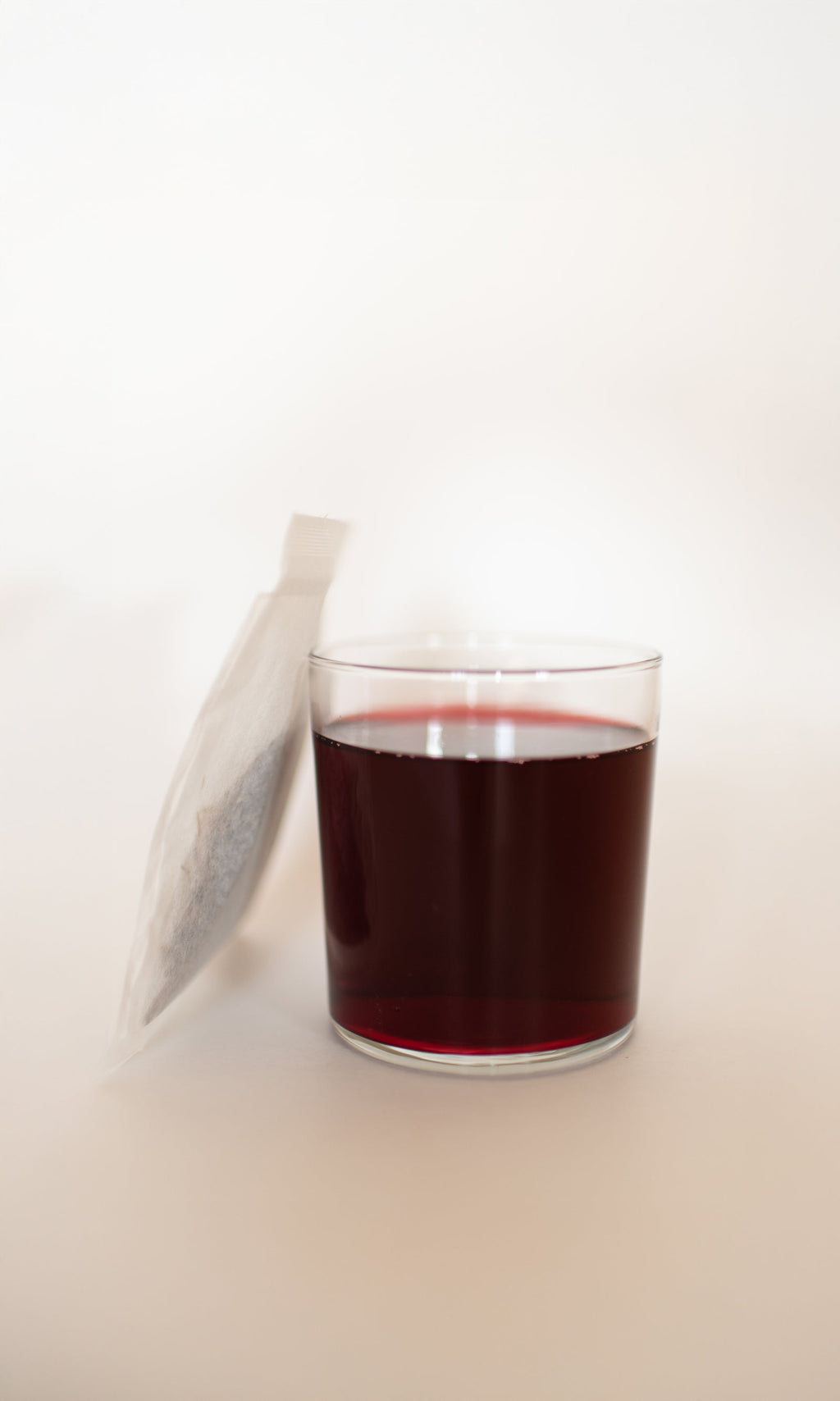 A clear glass of PLANTWALK COAST | Antioxidant Protection Herbal Infusion, a dark red drink, is placed next to a white tea bag on a light background.