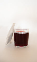 A clear glass of PLANTWALK COAST | Antioxidant Protection Herbal Infusion, a dark red drink, is placed next to a white tea bag on a light background.