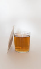 A clear glass of amber FIELD | Everyday Minerals Herbal Infusion by PLANTWALK sits on a white surface, steeped in mineral-rich water, with the tea bag standing upright beside it.