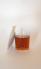 A clear glass of amber-hued FOREST | Healthy Hair & Skin Herbal Infusion by PLANTWALK sits on a light surface, tea bag resting upright—an inviting scene perfect for enjoying a boost of hair and nail strength.