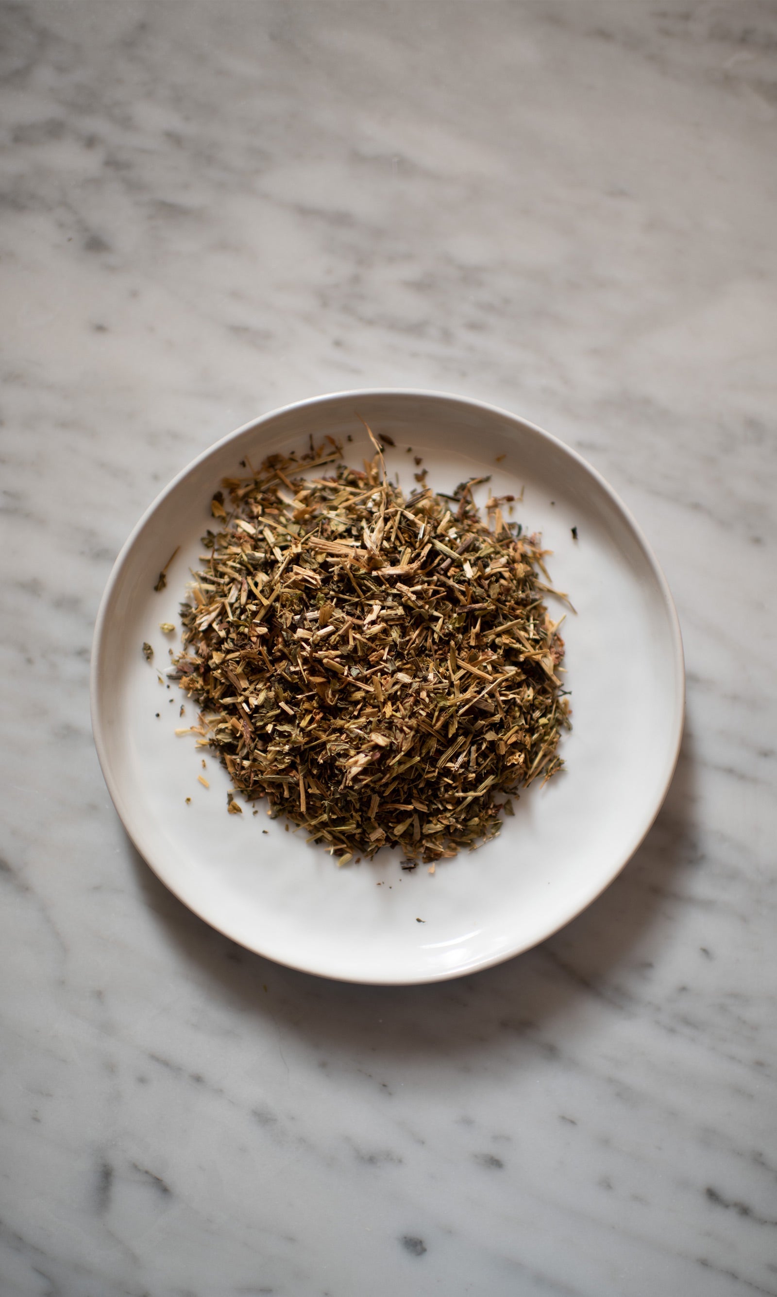 A white plate on a marble surface holds PLANTWALK’s RIVERBANK | Lymphatic Clarity Herbal Infusion, a blend of dried, shredded herbs commonly used for lymphatic system support and herbal lymphatic drainage.