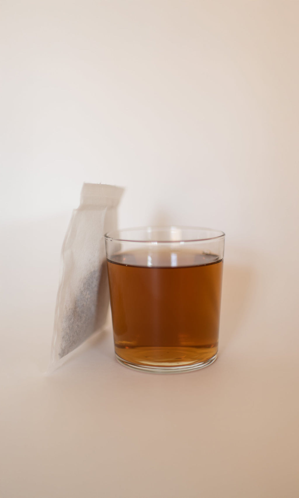 A glass of amber RIVERBANK | Lymphatic Clarity Herbal Infusion by PLANTWALK sits beside an unused tea bag on a light background, capturing the soothing ritual of supporting gentle lymphatic drainage.