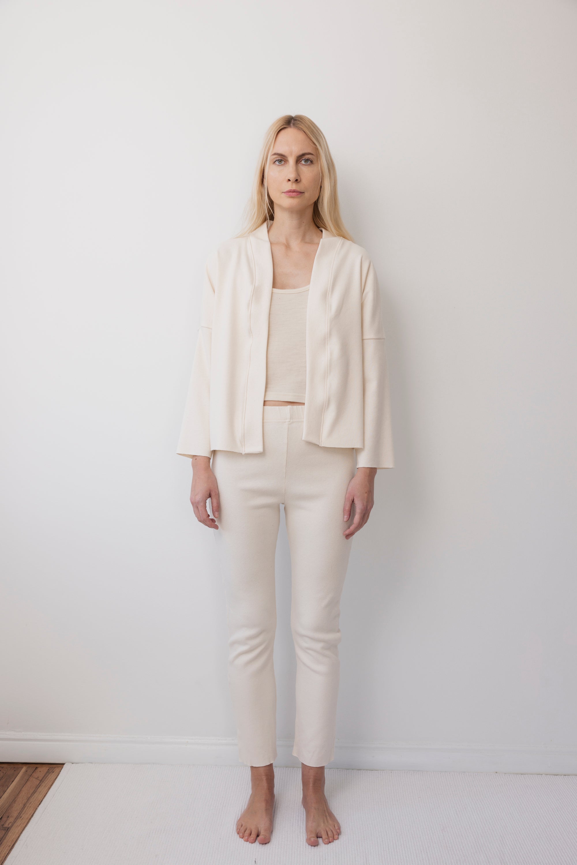 A woman with long blonde hair stands barefoot against a white wall, wearing the Wol Hide Easy Cardigan in Natural as part of a minimalist cream outfit with a matching top and slim pants.
