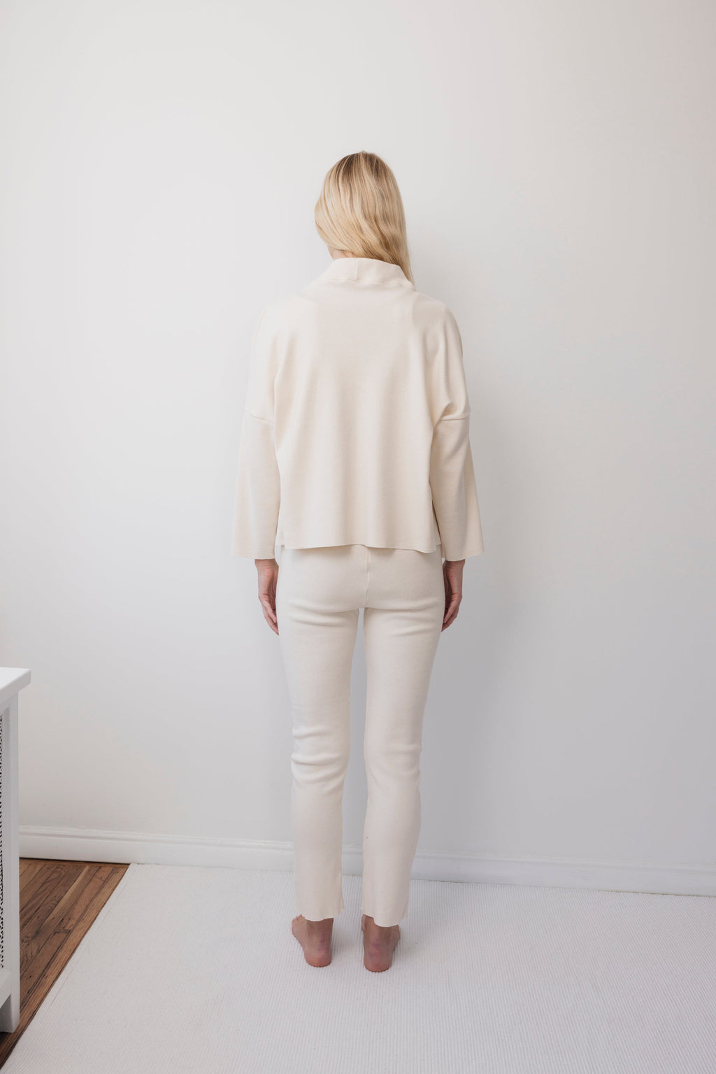 A person with long blonde hair stands barefoot in a minimalist white room, wearing the Wol Hide Easy Cardigan in Natural with fitted pants.