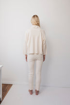 A person with long blonde hair stands barefoot in a minimalist white room, wearing the Wol Hide Easy Cardigan in Natural with fitted pants.