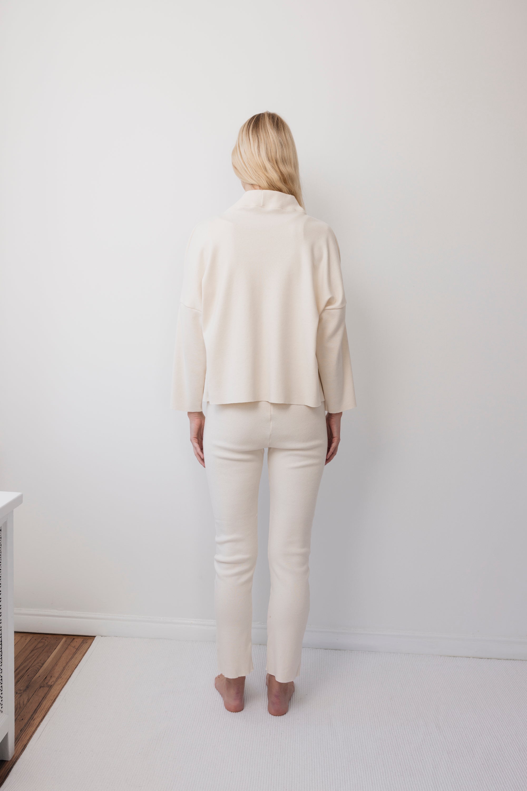 A person with long blonde hair stands barefoot in a minimalist white room, wearing the Wol Hide Easy Cardigan in Natural with fitted pants.