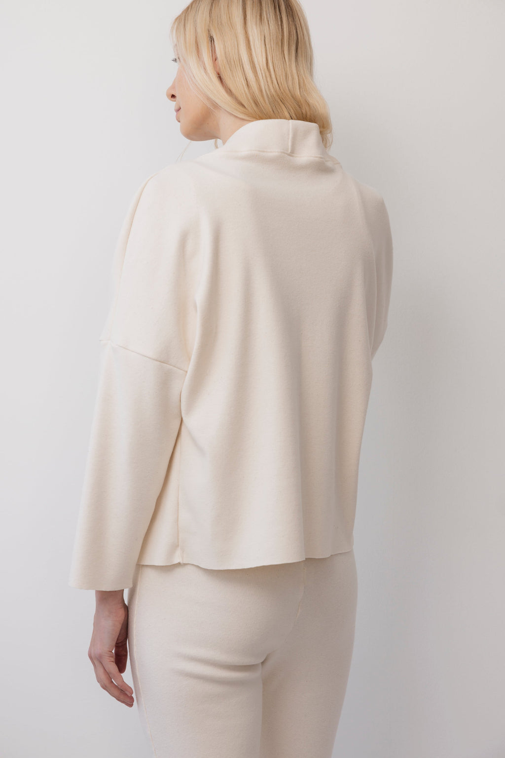 A person with light blonde hair is shown from the back, wearing the Wol Hide Easy Cardigan in Natural, featuring a loose fit and wide rib collar, paired with matching pants. The background is plain white.