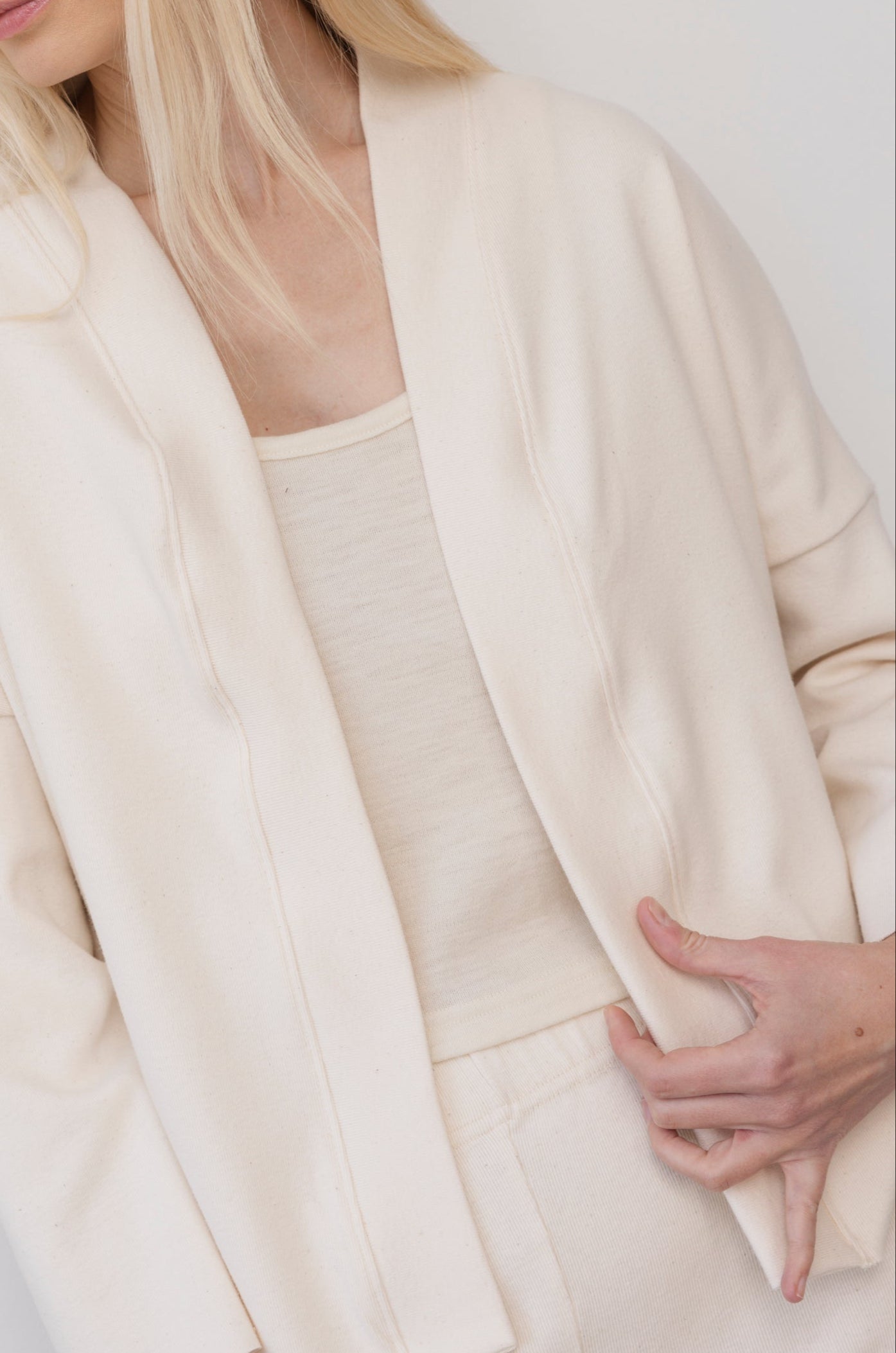 A person wearing Wol Hide’s Easy Cardigan in Natural stands against a plain background, lightly holding the edge of the cardigan. Their long blonde hair falls over one shoulder, and they are styled with matching pieces for a soft, cohesive look.