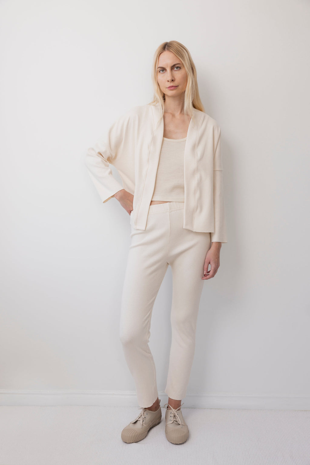A woman with long blonde hair stands against a white wall, wearing the Wol Hide Easy Cardigan in Natural with matching top and fitted pants, plus beige sneakers. She looks at the camera, relaxed, with her hands by her sides.
