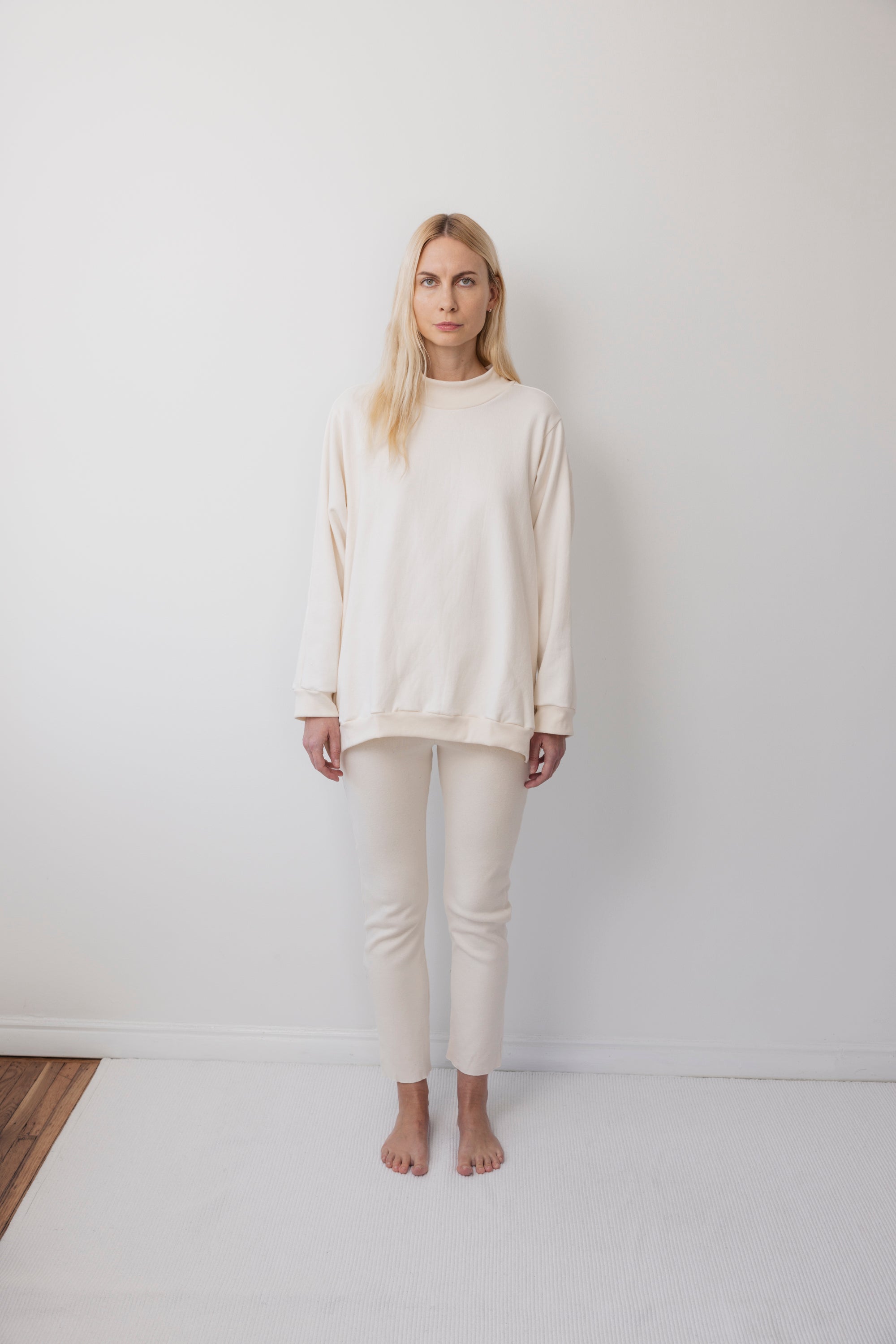A person with long blonde hair stands barefoot against a plain white wall, wearing the Wol Hide Big Sweatshirt in Natural with matching fitted pants, creating a minimal, simple look.